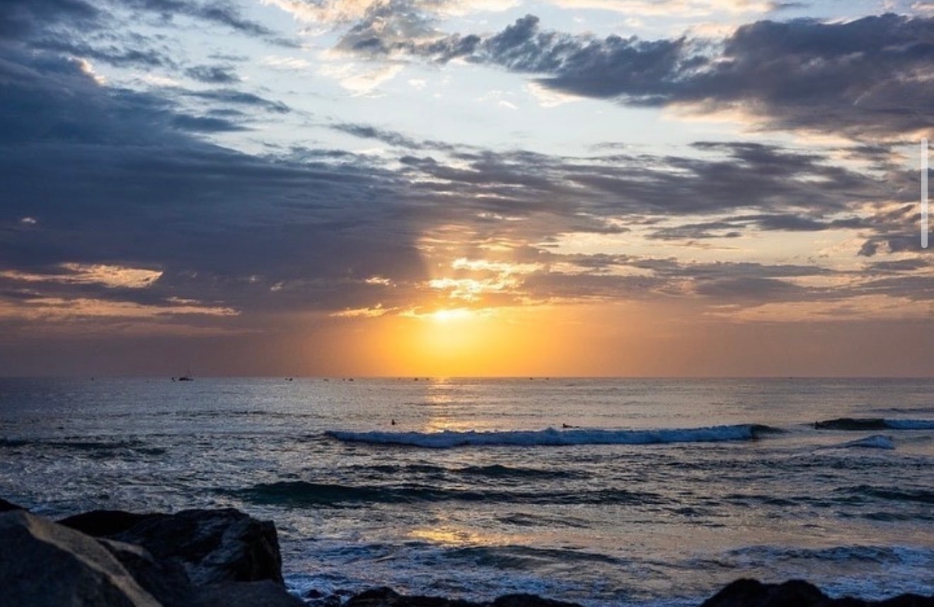 The sun dropping through a break in heavy cloud over a reeling point break, rocks in the foreground.