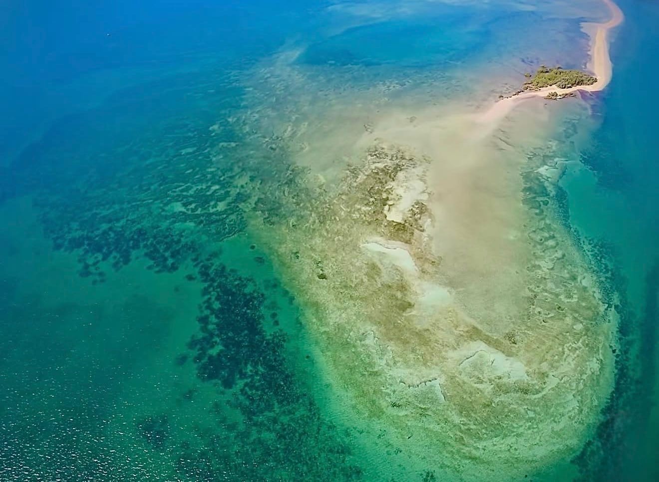 An aerial frame of a sand spit and reef, jade shallows shading into deep blue at the edges.