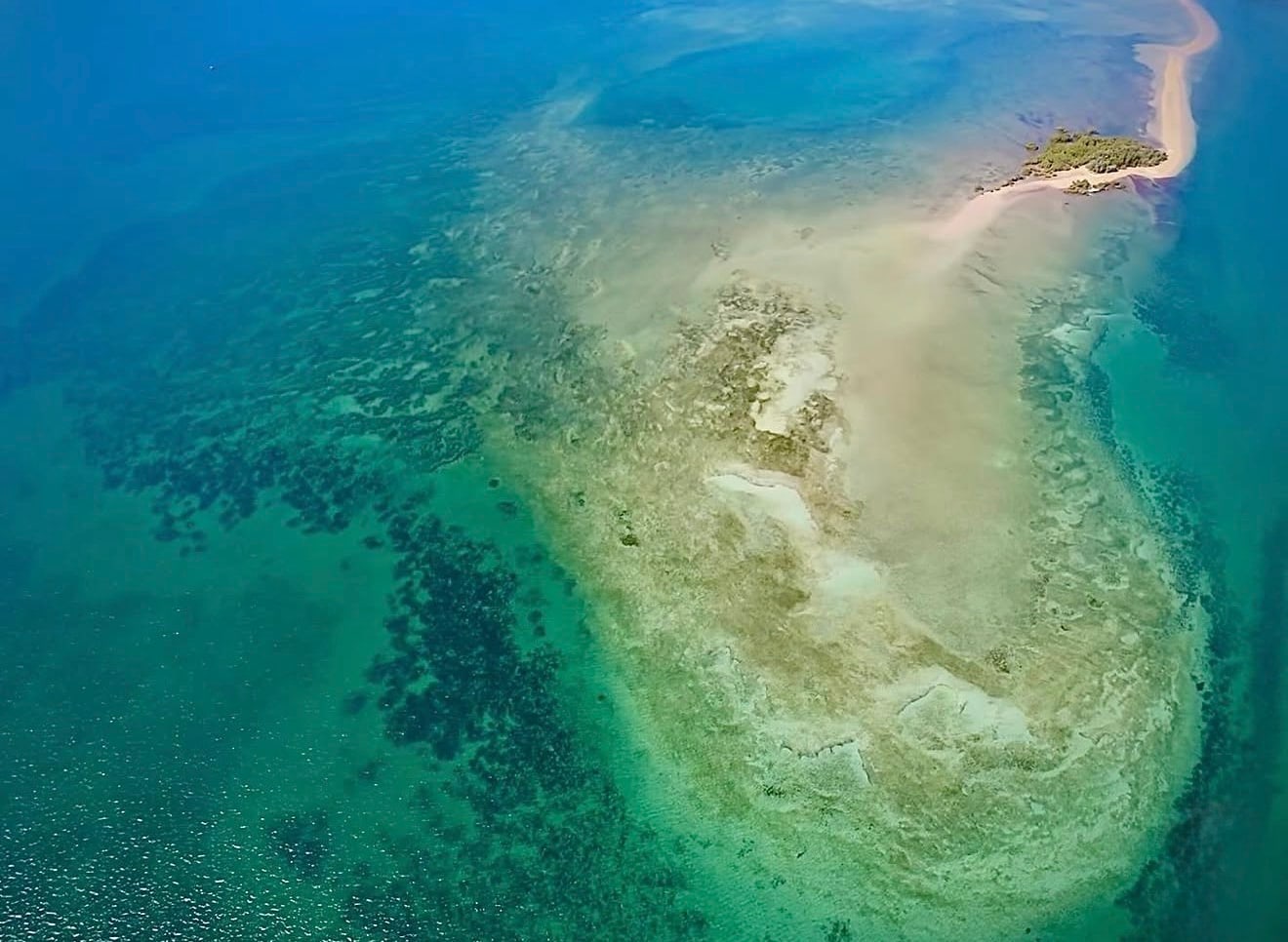 An aerial frame of a sand spit and reef, jade shallows shading into deep blue at the edges.